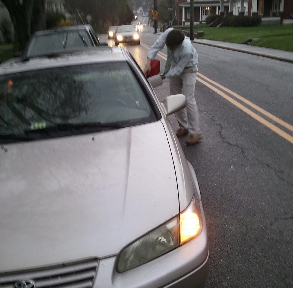 A Lampoon staffer in shirt and tie refuels a sedan by hand from a red gas can on the shoulder of a wet two-lane road at dawn.